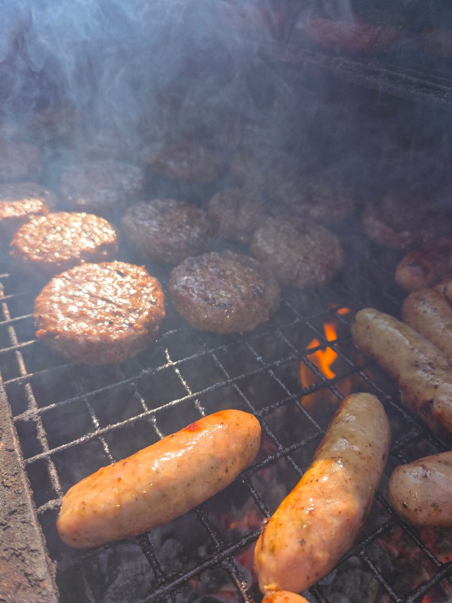 Sausages and burgers sizzle on the grill over hot coals, with smoke rising from the delicious BBQ