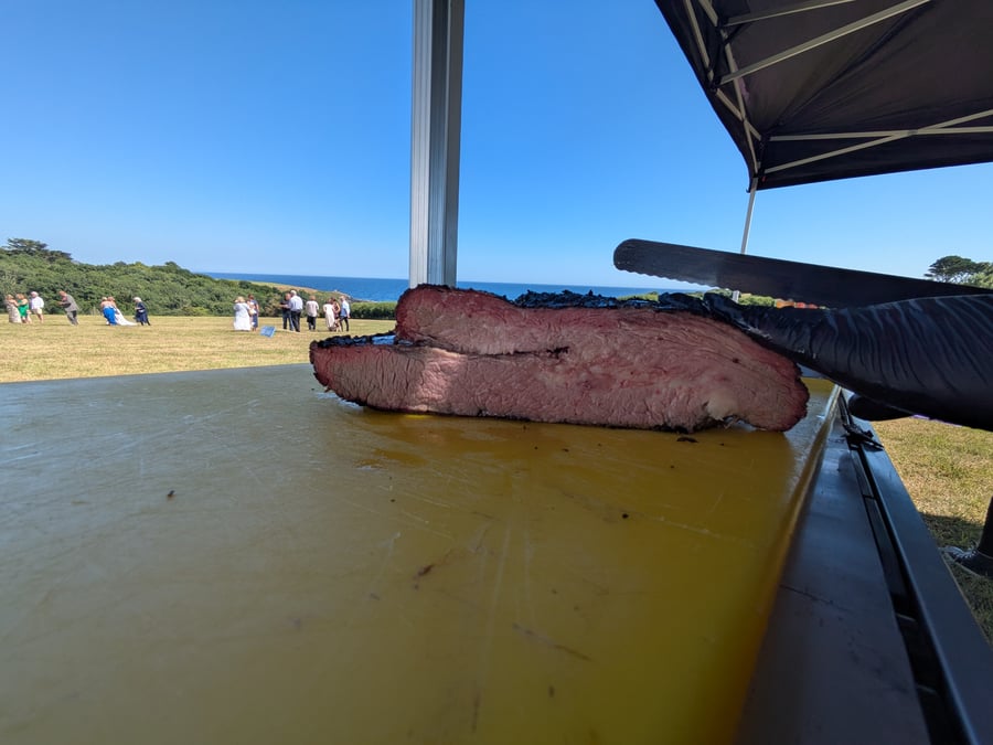 A chef carves succulent smoked brisket on a yellow cutting board at an outdoor event overlooking the sea