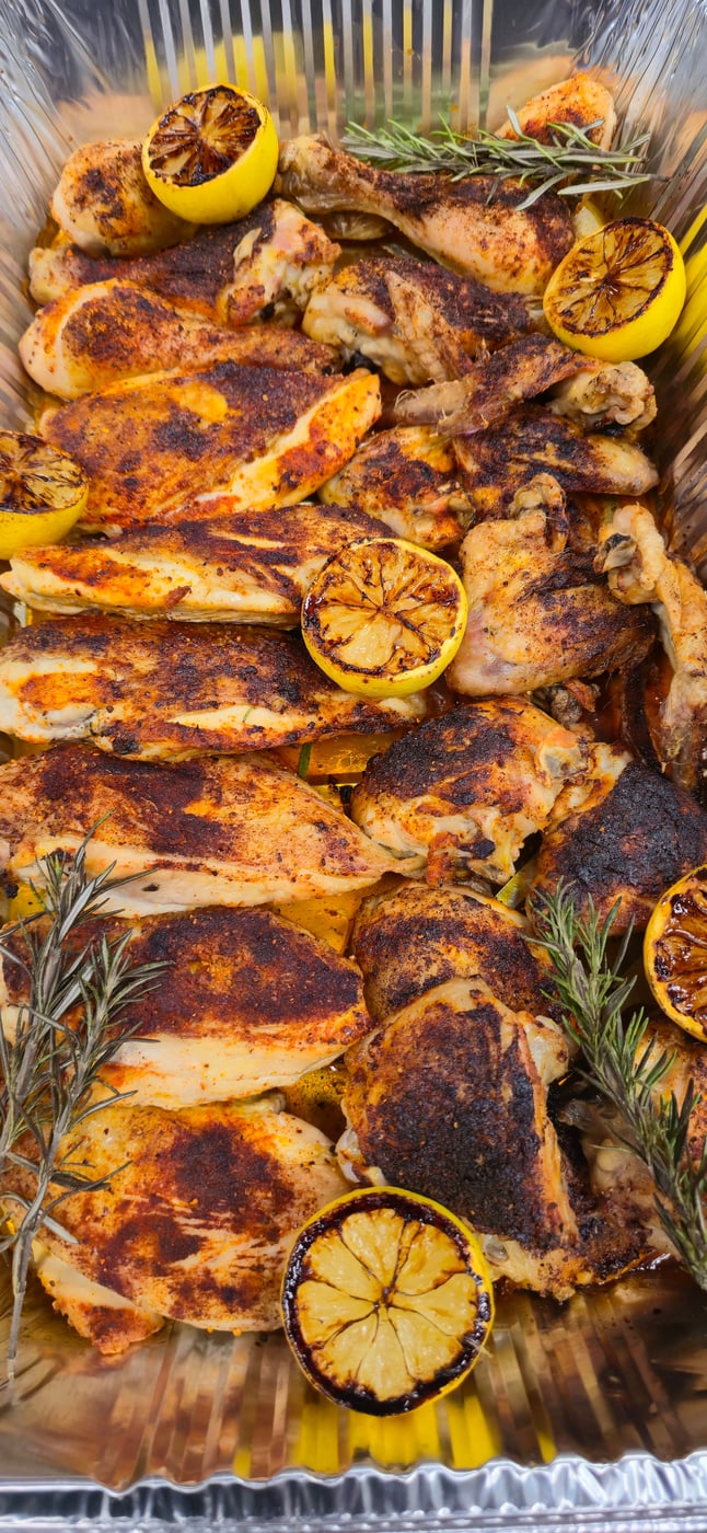 A foil tray filled with succulent grilled chicken pieces, garnished with charred lemon halves and fresh rosemary sprigs