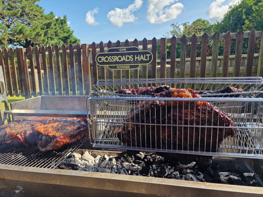 Large cuts of meat are slow-cooked over hot coals on a rotisserie grill at Crossroads Halt, Scorrier