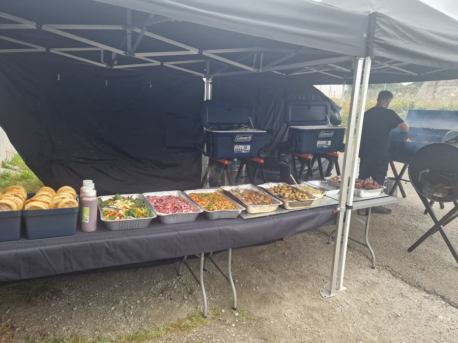 A chef cooks on a large BBQ grill at a film or TV unit base, with a buffet of salads, potatoes, and bread rolls laid out under a gazebo
