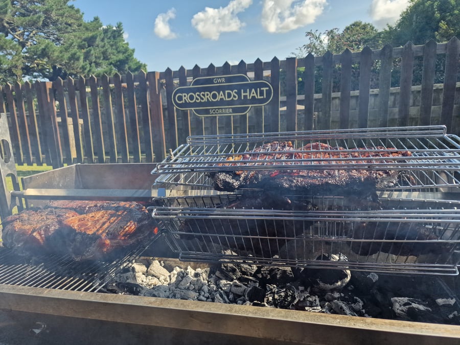 Large cuts of meat are slow-cooked over hot coals on a custom-built BBQ grill, with a railway sign in the background