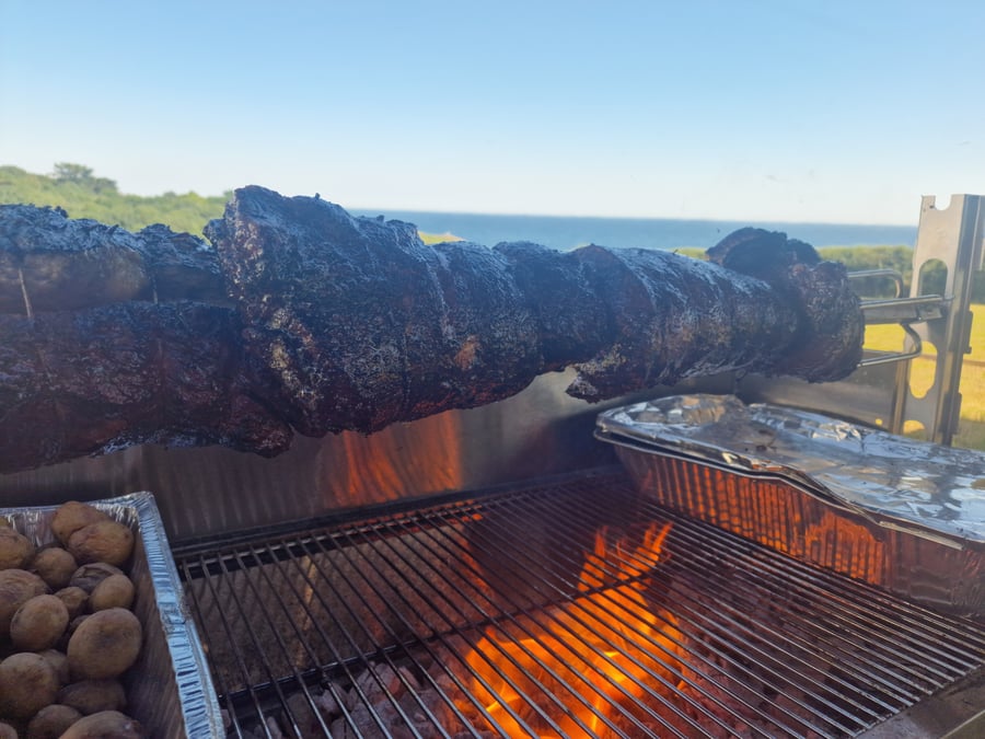 A hog roast slowly cooking over an open fire with roasted potatoes, set against a beautiful coastal backdrop