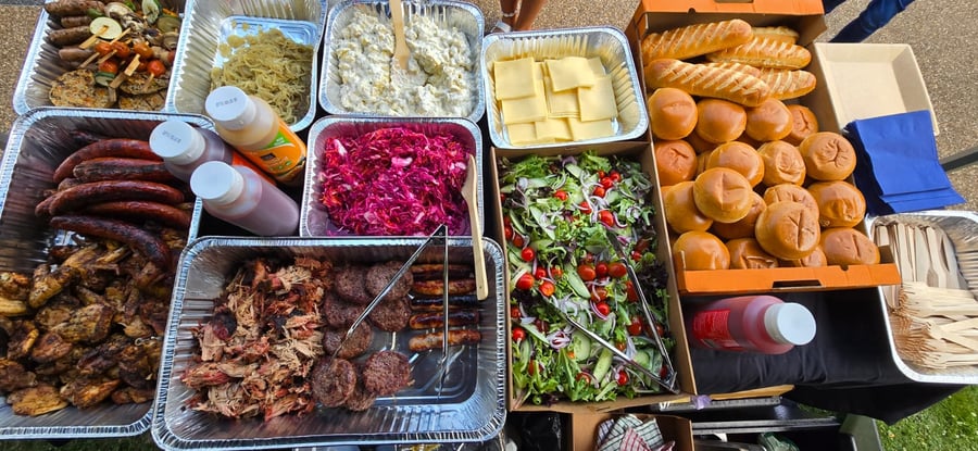 An overhead view of a buffet spread featuring pulled pork, sausages, burgers, salads, and bread rolls