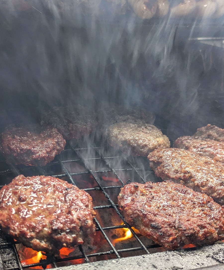 Juicy burgers sizzle on the grill, with smoke rising from the hot coals, promising a delicious BBQ feast