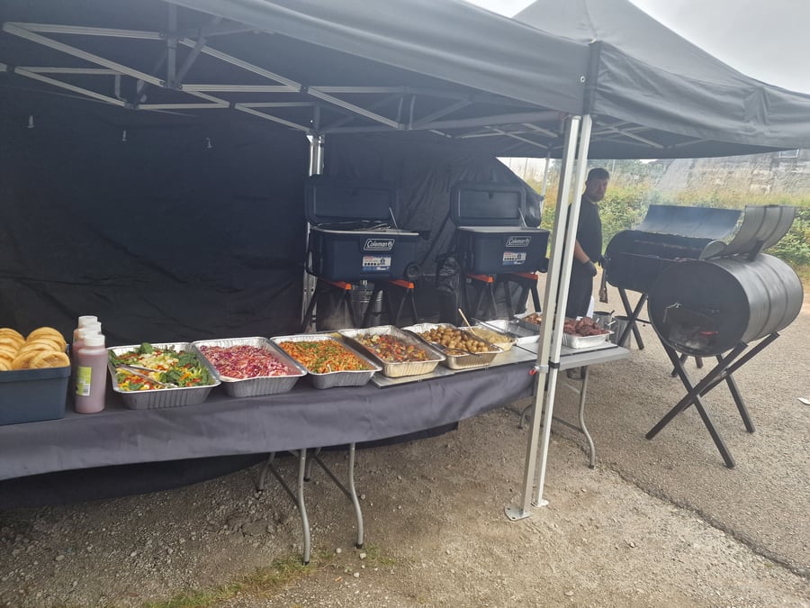 A chef tends to a large BBQ smoker next to a buffet table laden with salads, rolls, and hot dishes under a black gazebo