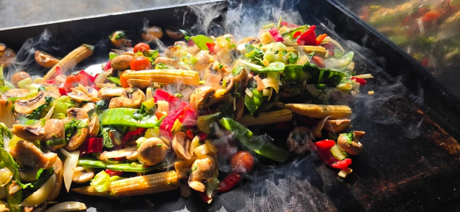 Steaming vegetables, including mushrooms, baby corn, and peppers, cooking on a large griddle