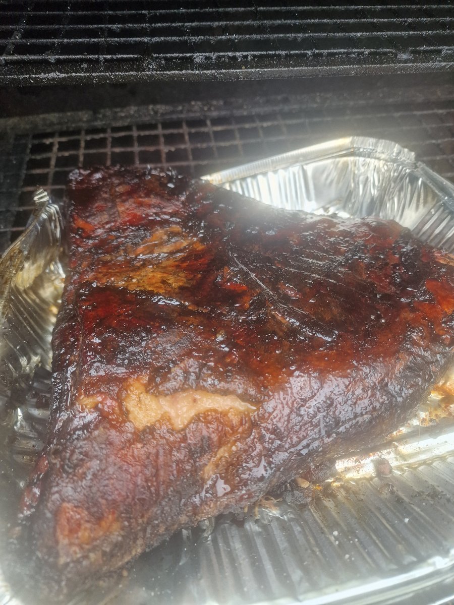 A large piece of slow-cooked, glazed meat rests in a foil tray on a BBQ grill, ready for carving