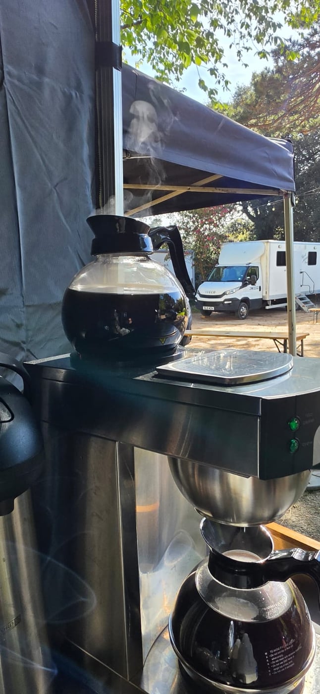 Freshly brewed coffee steaming on a catering unit base, with a production trailer and van in the background
