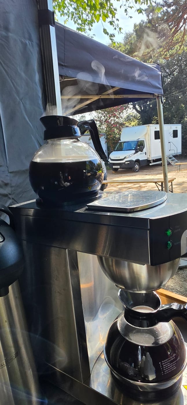Freshly brewed coffee steaming under a gazebo at a film or TV production unit base, with crew vehicles in the background