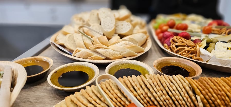 A buffet spread featuring sliced baguette, crackers, olive oil and balsamic vinegar, and a platter of cheeses, fruit, and pretzels