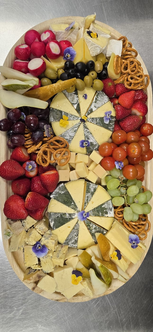 A vibrant cheese board featuring various cheeses, fresh fruit, radishes, olives, and pretzels, garnished with edible flowers (2)