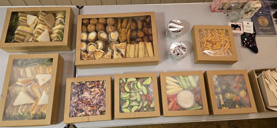 A variety of buffet food in individual boxes, including sandwiches, scotch eggs, quiche, pasta, salads, and fruit