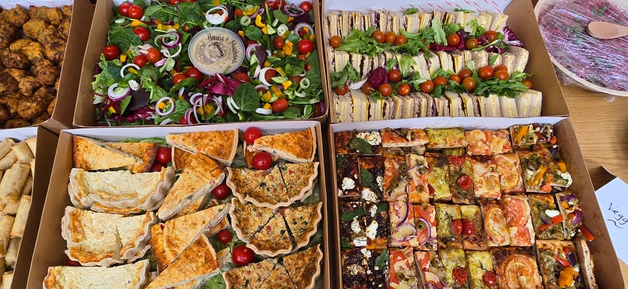 A vibrant selection of buffet items, including fresh salad, sandwiches, quiches, and focaccia, presented in catering boxes