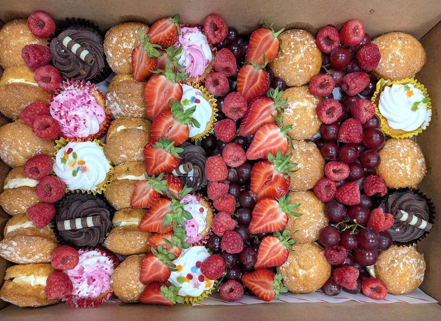 A vibrant dessert buffet box featuring mini cupcakes, cream-filled choux buns, fresh strawberries, raspberries, and grapes