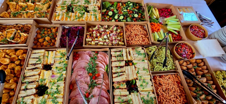 An overhead view of a large buffet spread featuring sandwiches, salads, pasta, and various savoury dishes in cardboard boxes