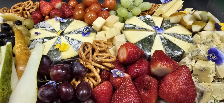 A vibrant cheese and fruit platter featuring various cheeses, fresh strawberries, grapes, pears, olives, and pretzels, garnished with