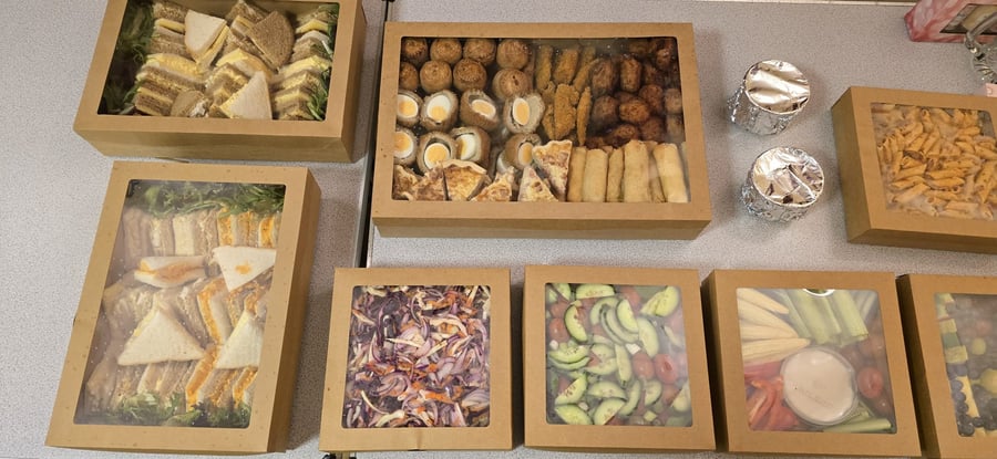 A selection of buffet food in cardboard boxes, including sandwiches, Scotch eggs, sausage rolls, pasta salad, and crudités