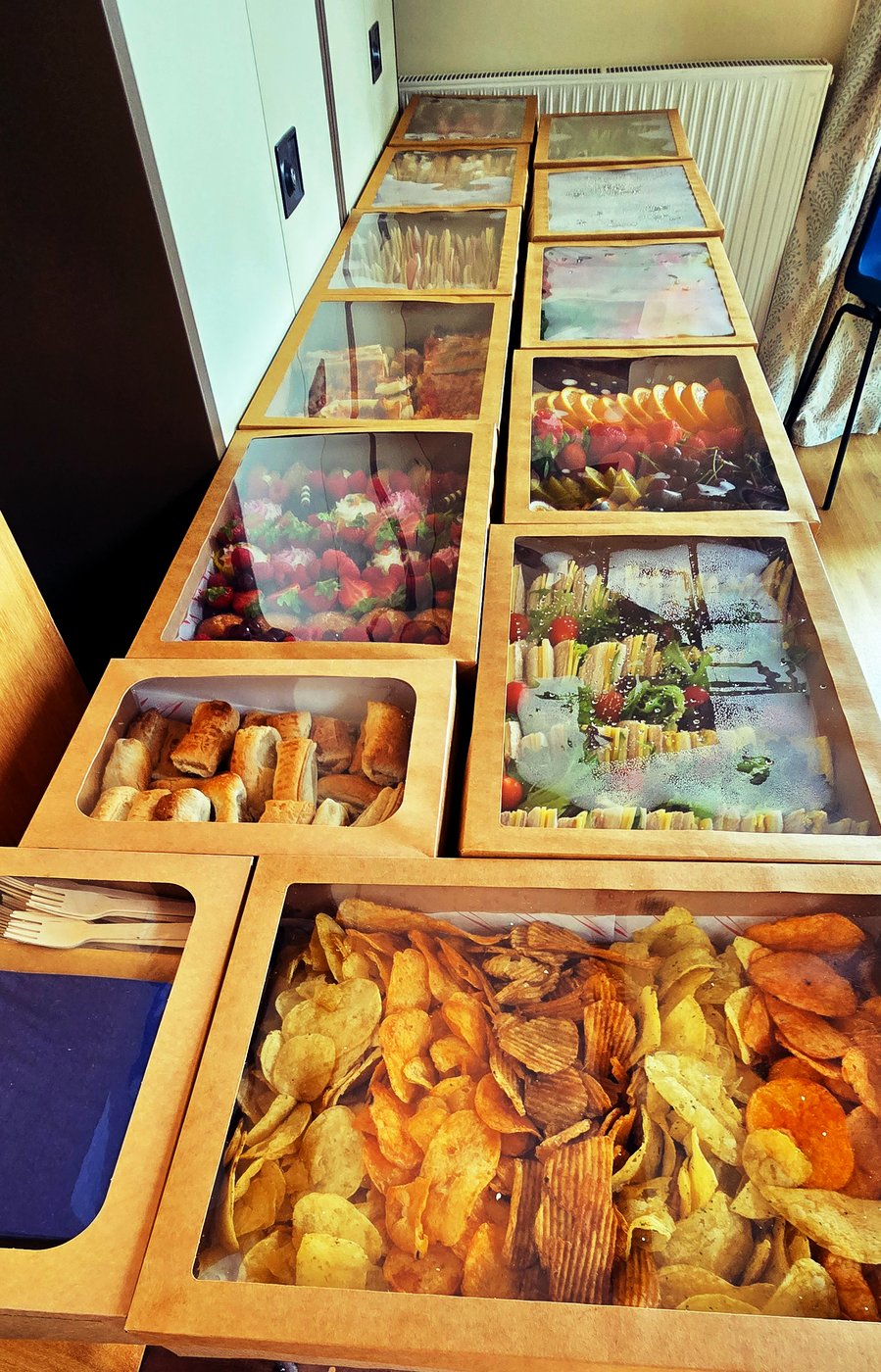 A long table laden with a variety of buffet boxes, including sausage rolls, sandwiches, crisps, fruit, and strawberries