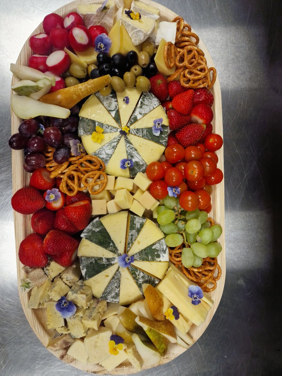 A vibrant cheese board featuring various cheeses, fresh fruit, radishes, olives, and pretzels, garnished with edible flowers