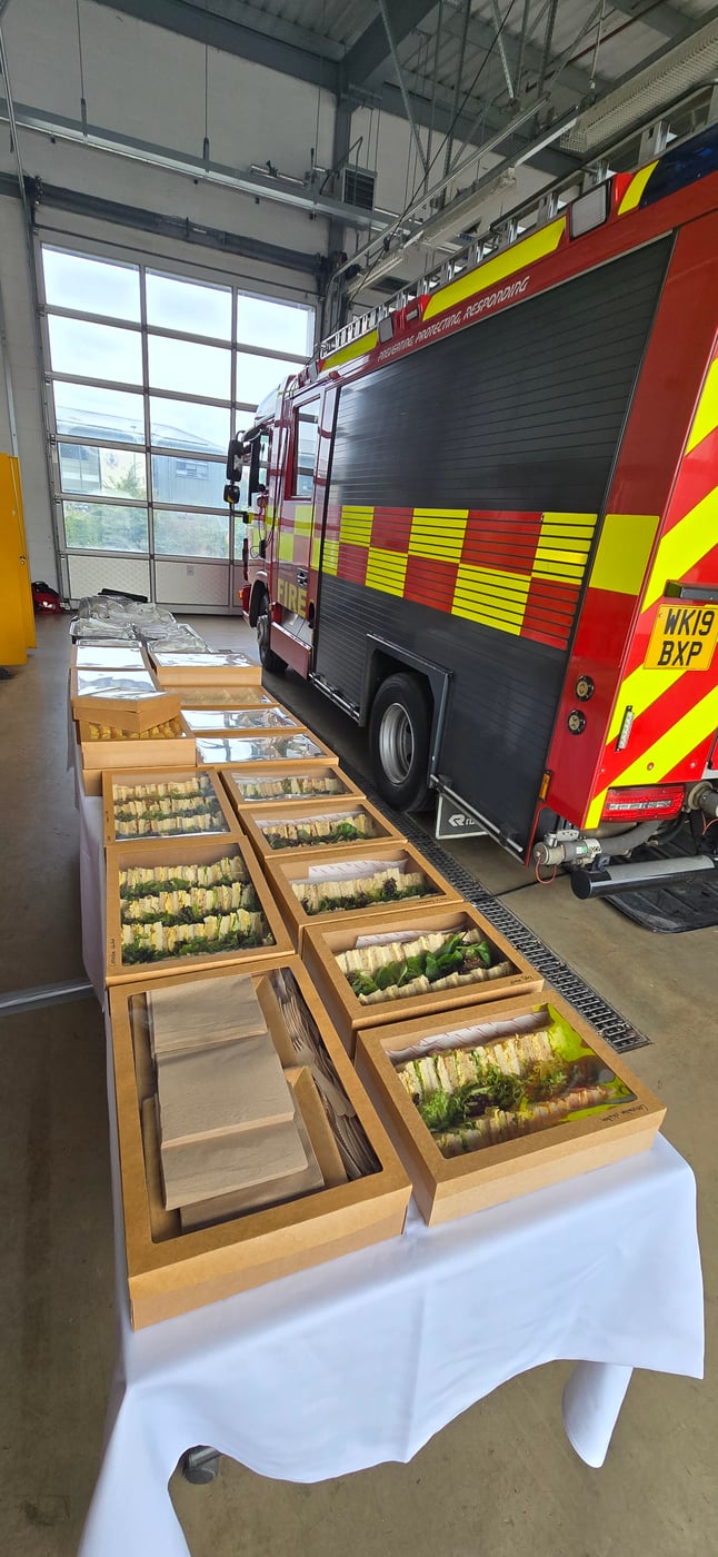 A table laden with individually boxed sandwiches and cutlery, set up for welfare catering next to a fire engine in a station