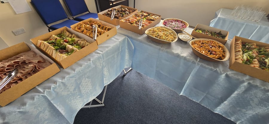 A varied buffet spread featuring sandwiches, cold meats, sausage rolls, and pasta salads, presented in eco-friendly packaging