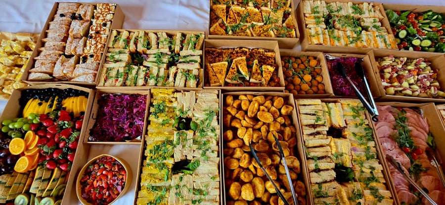 An overhead view of a large buffet spread featuring sandwiches, fruit platters, salads, and savoury pastries in cardboard boxes