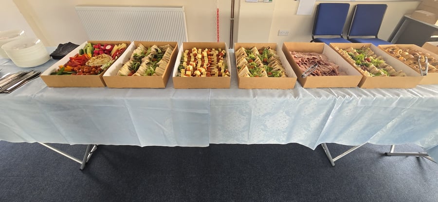 A long table laden with a variety of buffet food boxes, including sandwiches, crudités, cheese and pineapple sticks, and cold meats