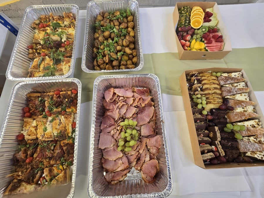 A varied buffet spread featuring quiches, roasted potatoes, sliced ham, a fresh fruit platter, and a dessert selection