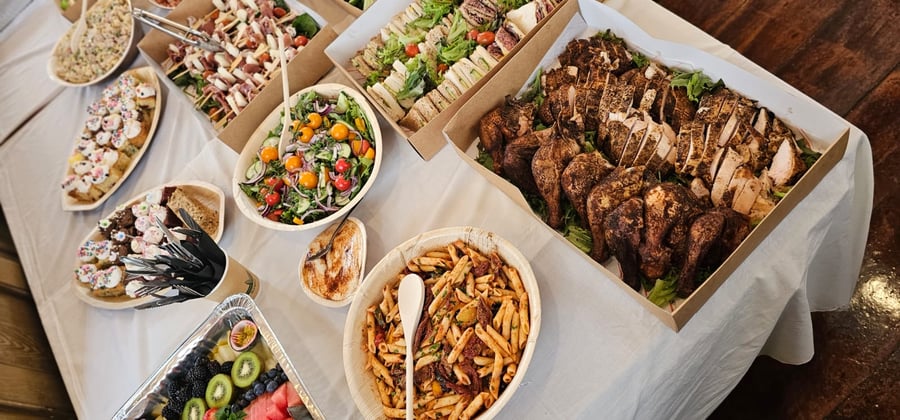A vibrant buffet spread featuring roasted chicken, pasta salad, fresh green salad, sandwiches, fruit, and sweet treats