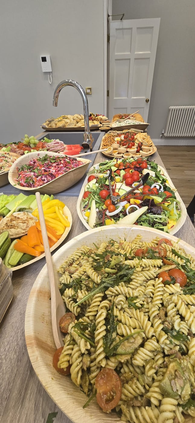 A vibrant buffet spread featuring pasta salad, mixed green salad, crudités with dip, and other savoury dishes