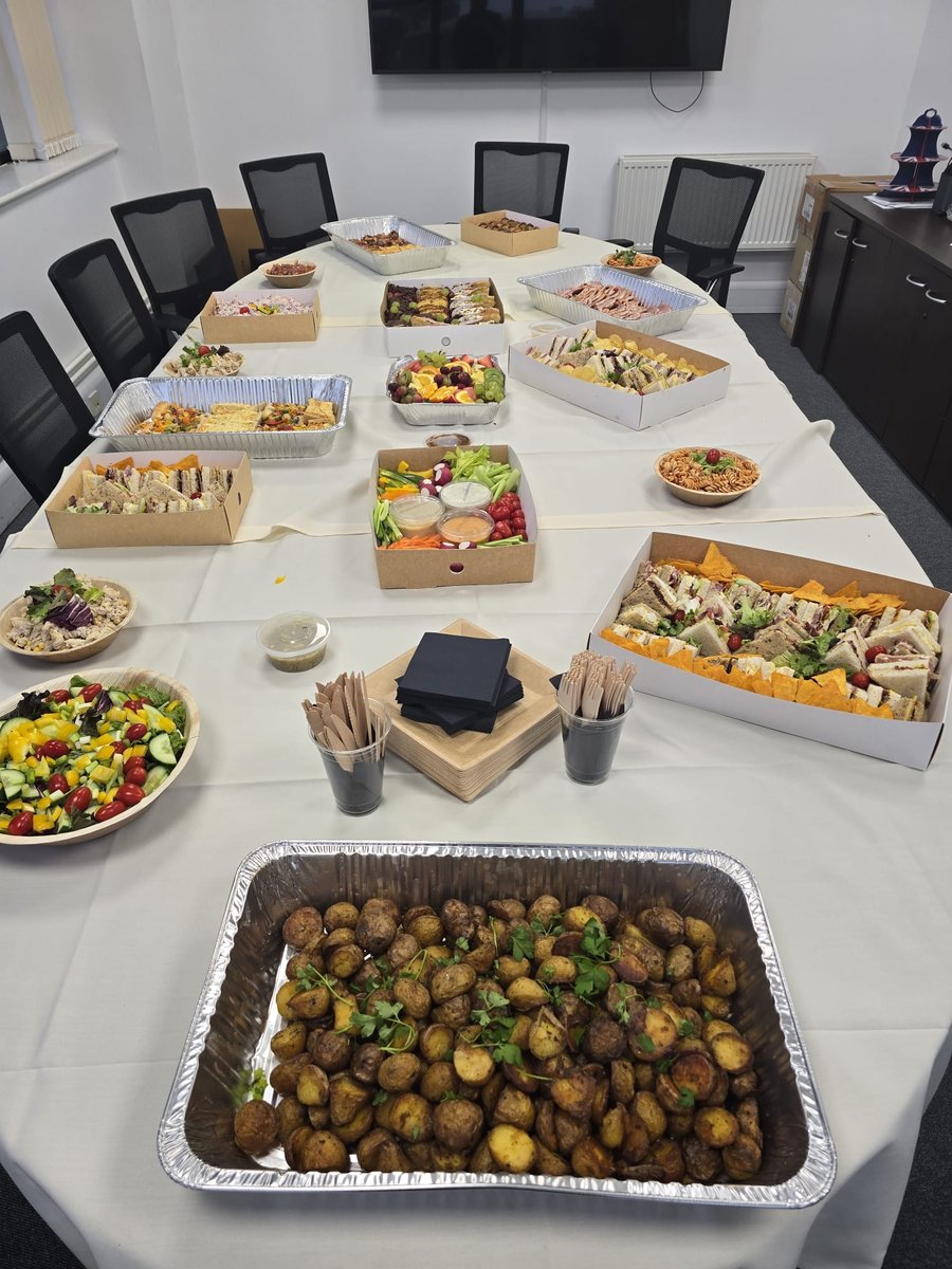 A conference room table laden with a variety of buffet dishes, including sandwiches, salads, fruit, and roasted potatoes
