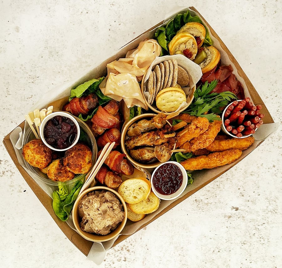 A delicious selection of buffet items, including pigs in blankets, chicken skewers, pâté, and crackers, presented in a catering box