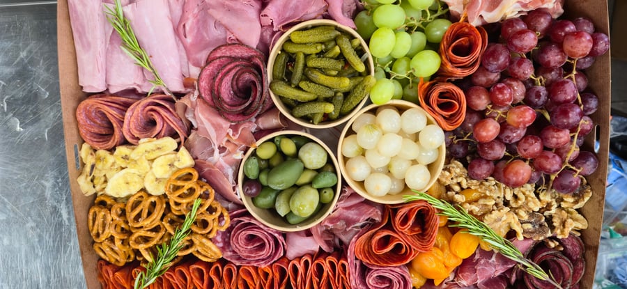 A vibrant buffet platter featuring cured meats, grapes, olives, cornichons, pickled onions, pretzels, and dried fruit