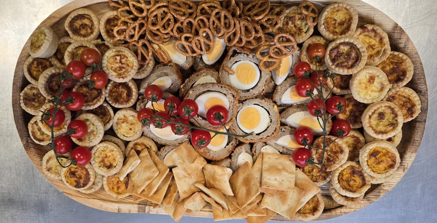 A wooden platter laden with mini quiches, Scotch eggs, pretzels, crackers, and cherry tomatoes, perfect for a buffet