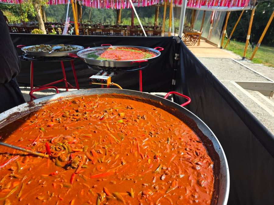 A large pan of paella with red peppers and other ingredients is cooking outdoors, with two smaller pans in the background