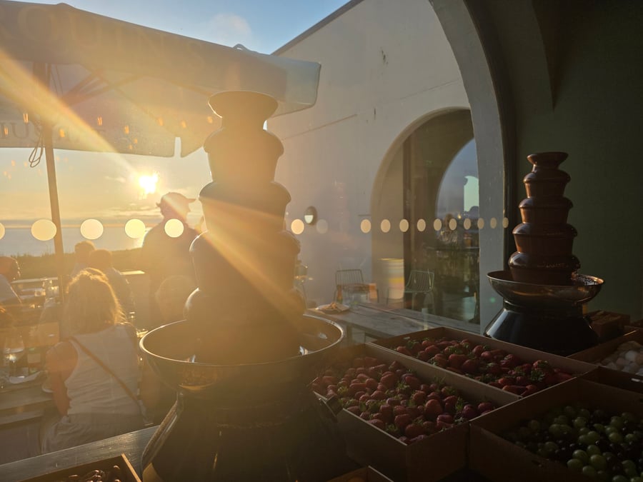 A chocolate fountain with fresh strawberries and grapes, set against a beautiful sunset over the sea