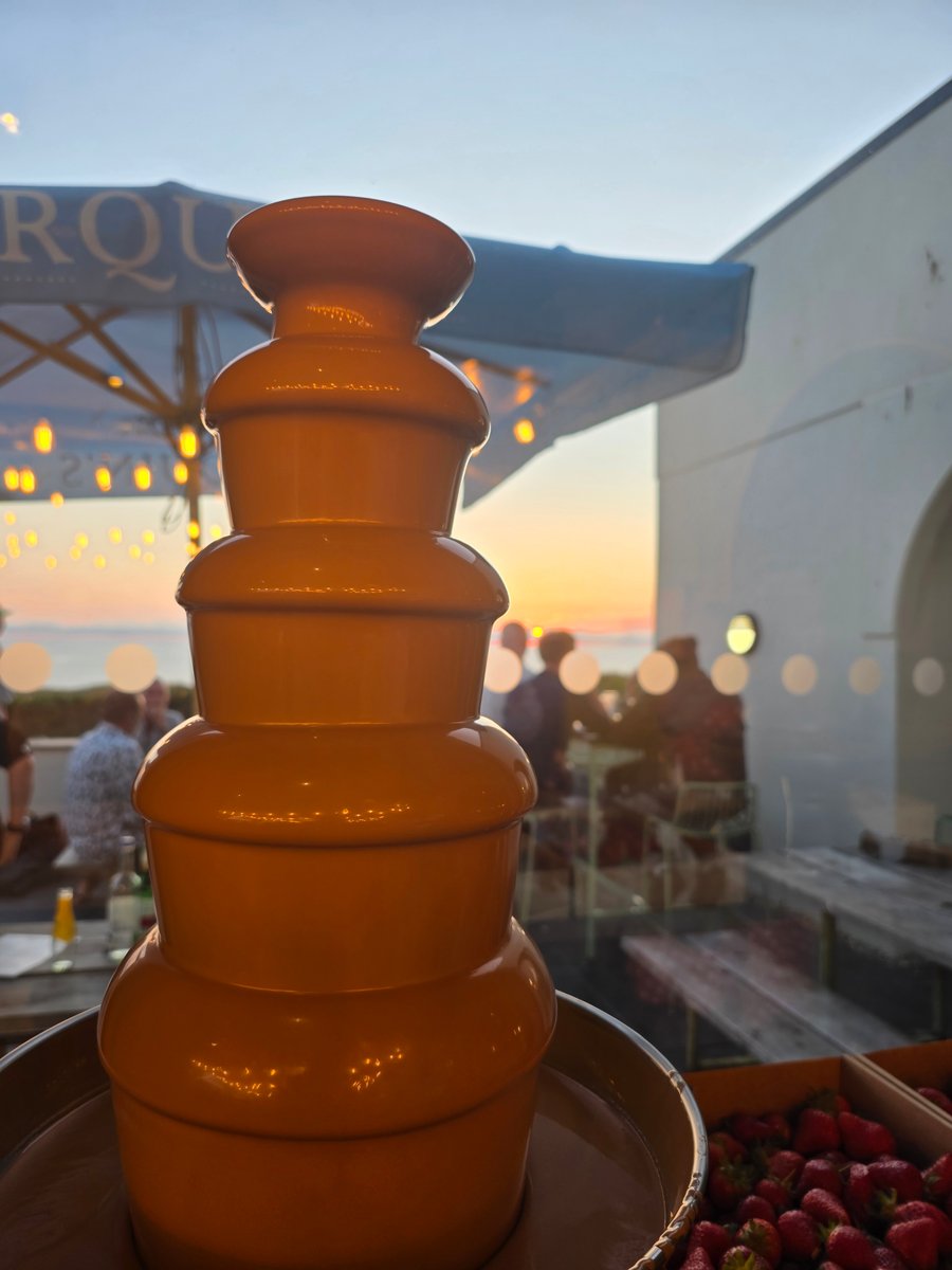 A chocolate fountain with fresh strawberries, set against a sunset backdrop at an outdoor event