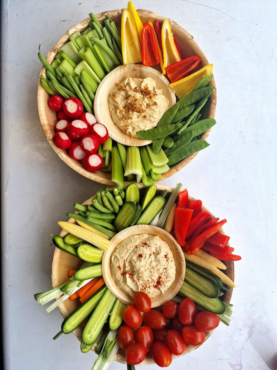 Two platters of fresh crudités and hummus are artfully arranged on palm leaf plates