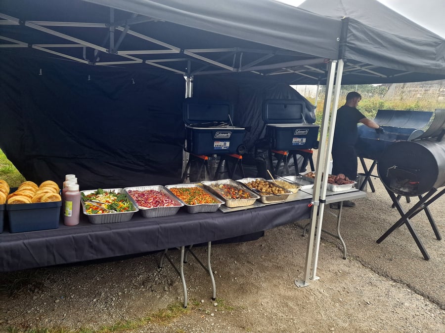 Outdoor catering setup with a buffet of salads and meats being cooked on a BBQ