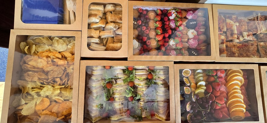 Assortment of catering boxes filled with sandwiches, fruit, pastries, crisps, and pizza
