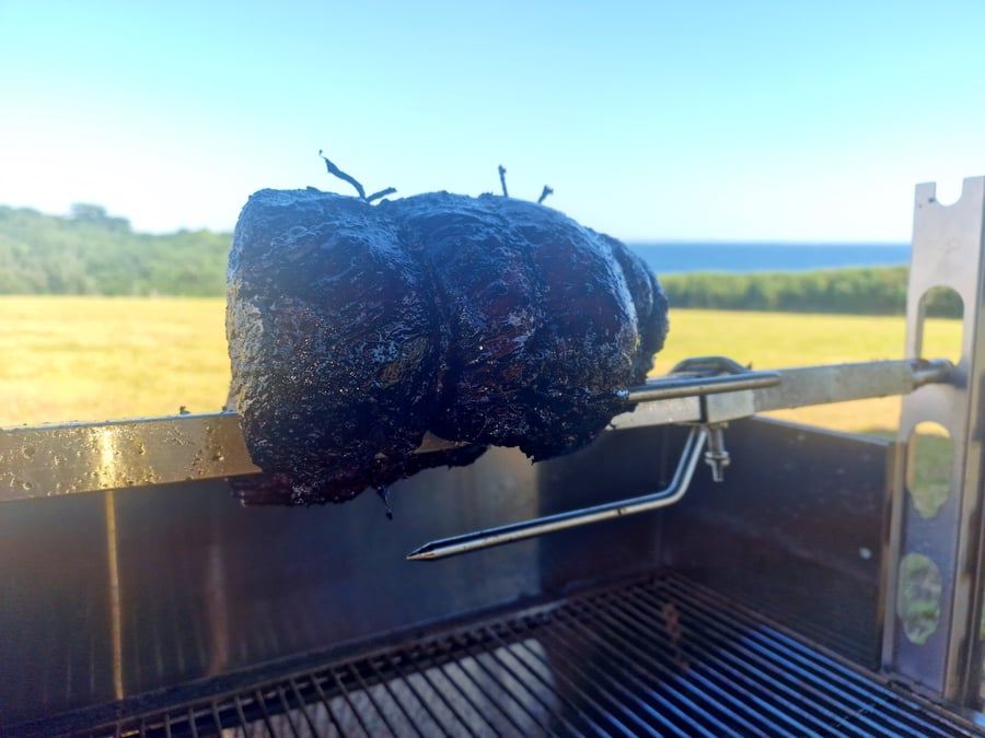 A large piece of meat is roasting on a spit outdoors with the sea in the background