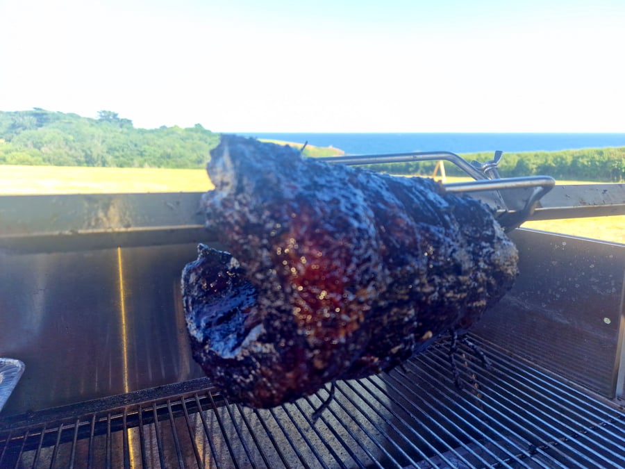 A large hog roast is slowly cooking on a spit with the sea in the background