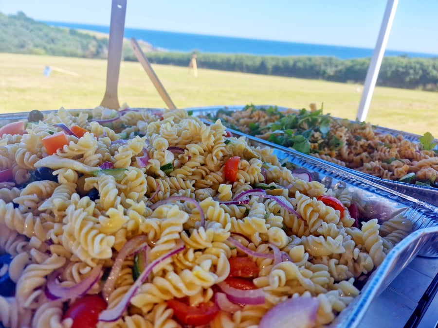 Two large trays of pasta salad are presented outdoors with a coastal view