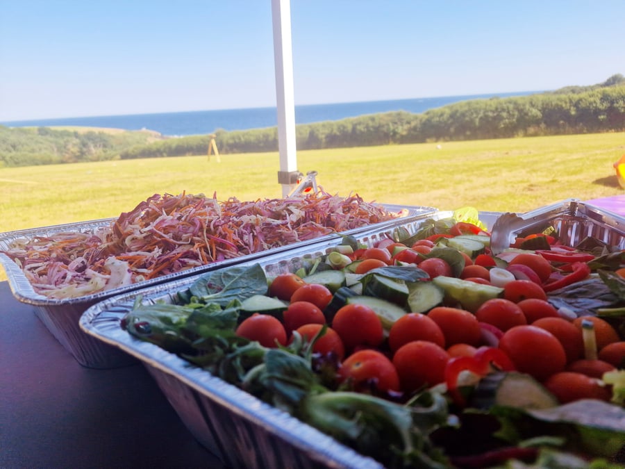 Two large catering trays of fresh salads and coleslaw are set up outdoors