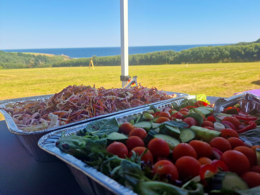 Two large catering trays of fresh salads sit on a table outdoors with a coastal view
