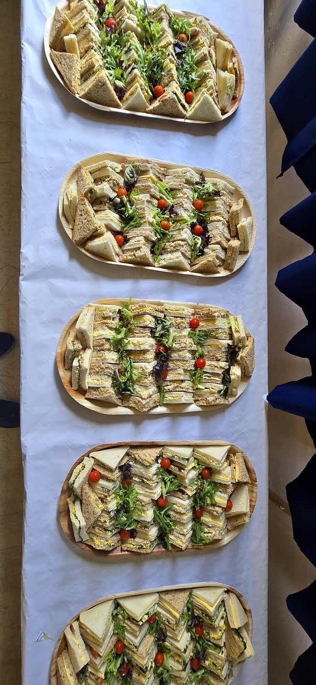 Five platters of assorted sandwiches are neatly arranged and garnished with greens and tomatoes