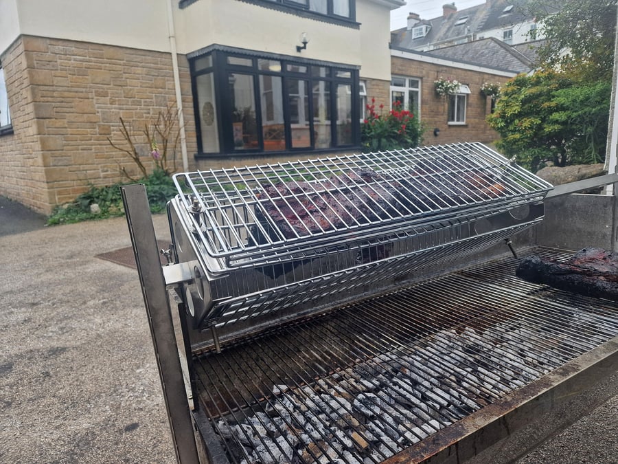 Meat is cooking on a large rotisserie grill outdoors with hot coals below