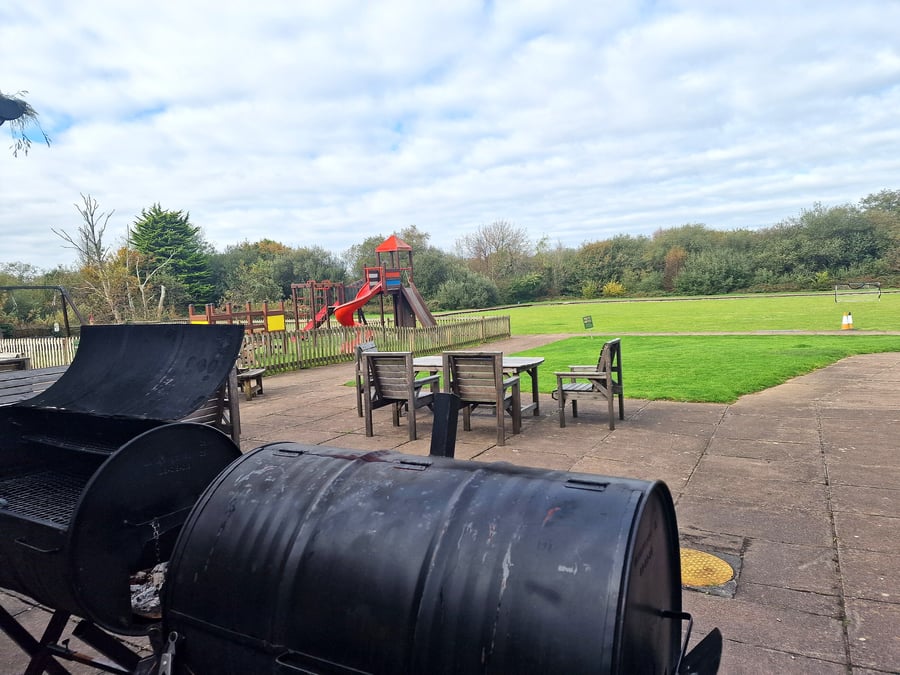 Two large barrel BBQs sit ready for cooking outdoors with a playground in the background