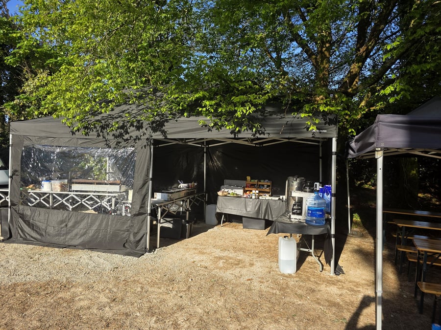 Outdoor catering setup with tents, tables, and food preparation equipment under trees